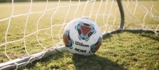 An image of a football in the corner of a football net, sitting on grass.